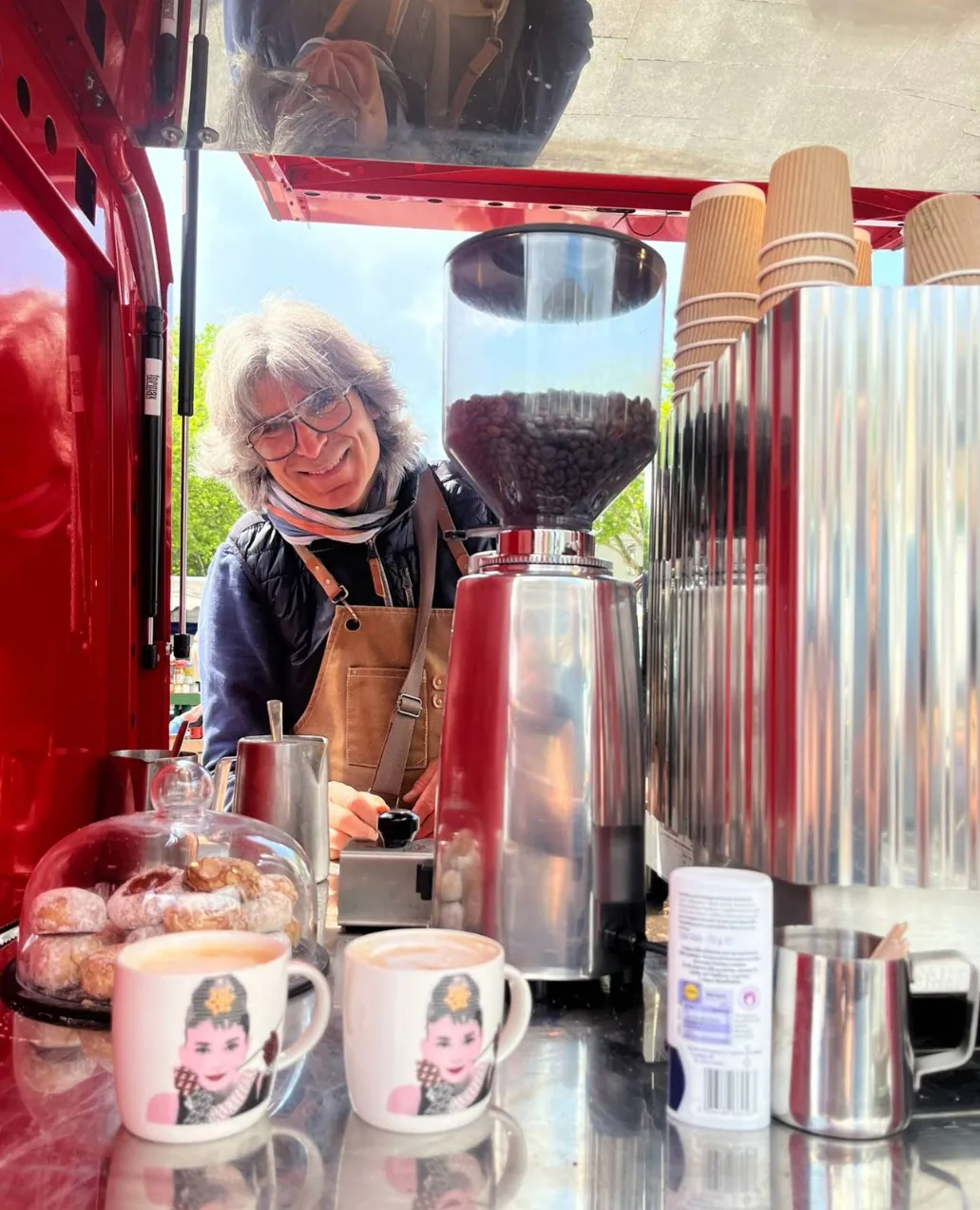 Lächelnde Barista in Schürze hinter der Theke der roten Ape, daneben Mühle mit Kaffeebohnen und zwei Cappuccino