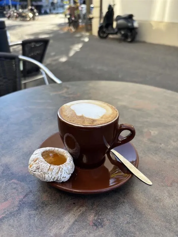 Cappuccino in einer braunen Tasse mit Gebäck auf einem Tisch vor dem Caffe-Caico, im Hintergrund die Straße und ein Motorroller