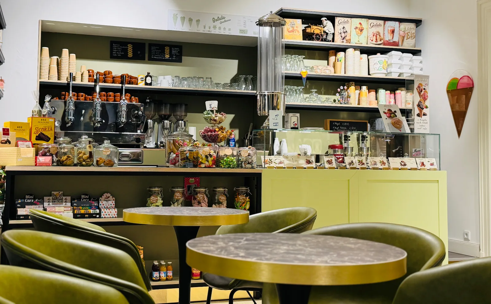 Gastraum im Caffe-Caico: runde Bistrotische mit olivgrünen Sesseln vor Eistheke und verchromter Hebel-Espressomaschine, dahinter Regale mit Bechern und Gläsern